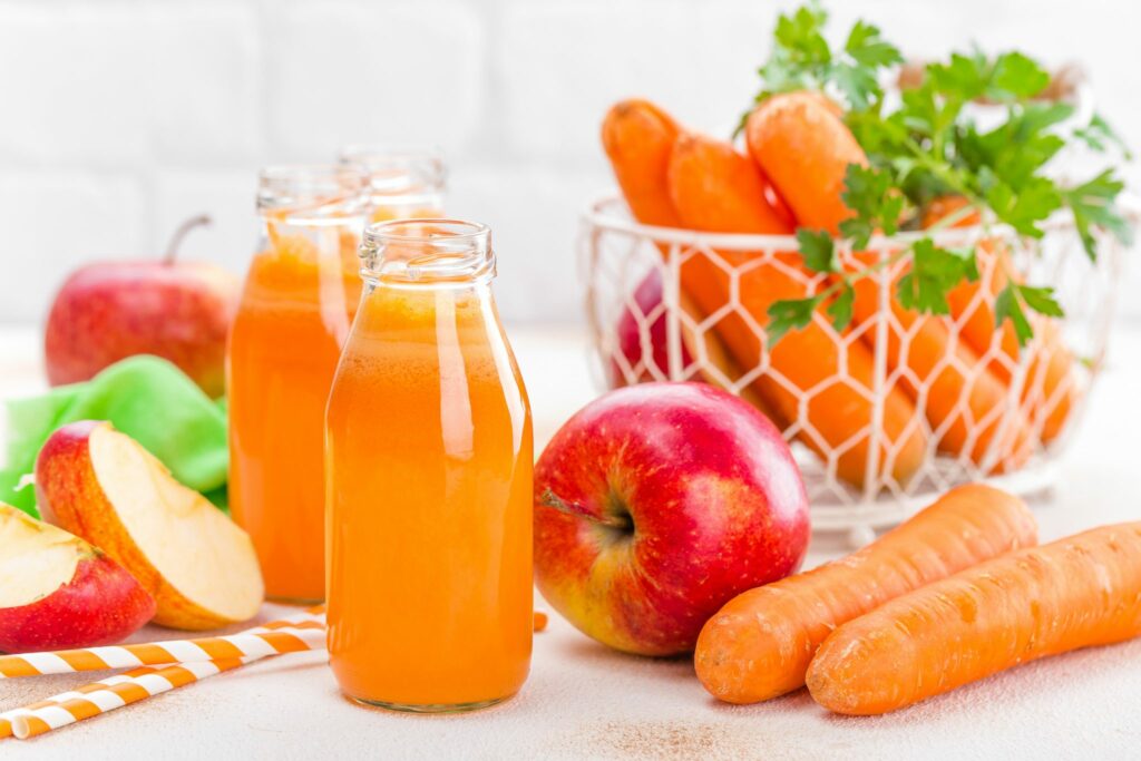 Três garrafinhas com suco na cor laranja. Ao lado, estão dispostas maças inteiras e cortadas, cenouras sobre a mesa e um cesto com cenouras e sasinhas.
