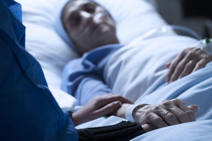 Pessoa dormindo em cama de hospital, e outra pessoa repousa a mão em seu braço.