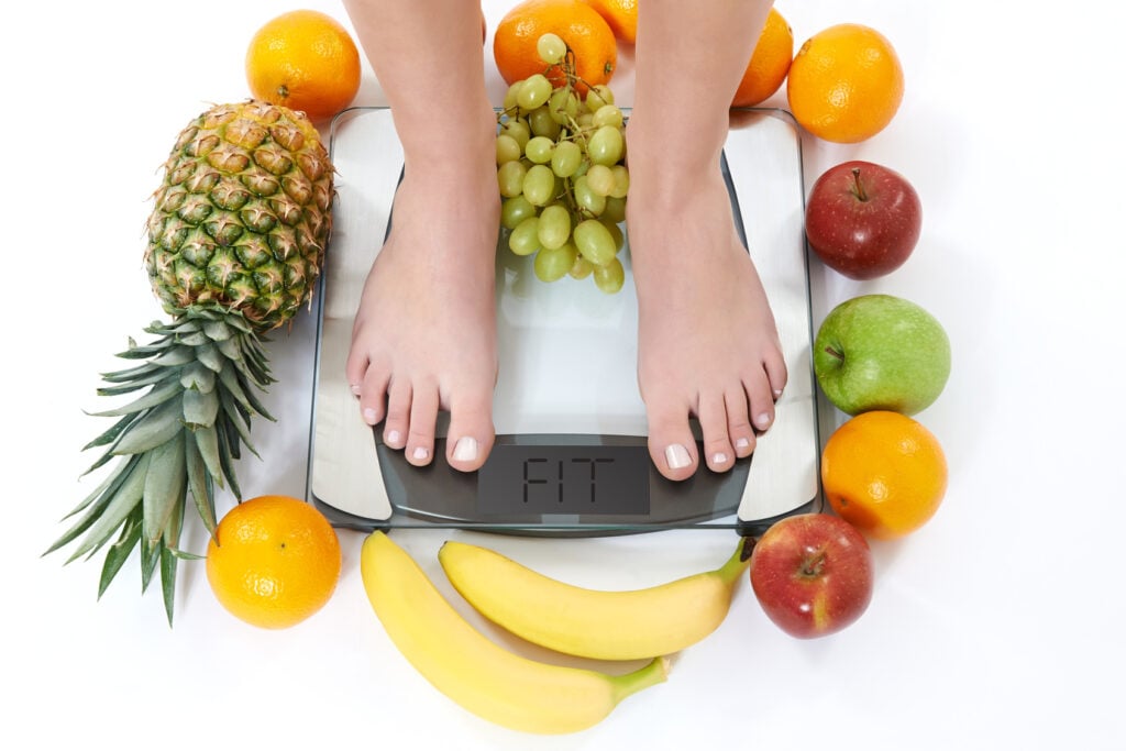 Pés femininos sobre uma balança de peso na cor branca. Em volta da balança, vários tipos de frutas como banana, laranja, abacaxi, uva e maças.
