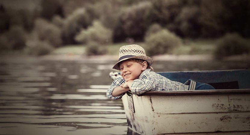 Imagem de um lago com um barquinho. Nesse barquinho um menino pensativo. Ele está vestindo uma camisa xadrez e um chapéu de palha.
