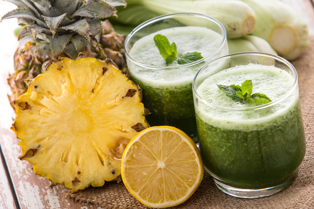 Dois copos cheios de suco de couve com abacaxi. Ao lado dos copos um abacaxi cortado ao meio. Os copos estão decorado com folhas de hortelã.
