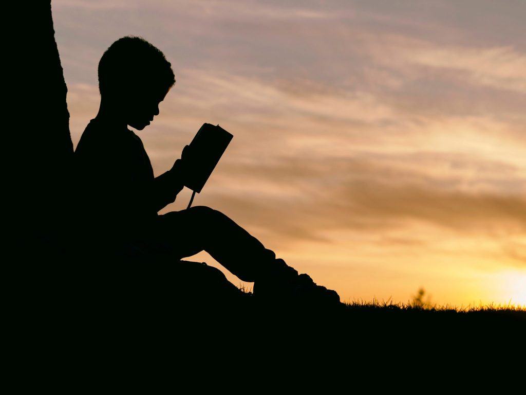 Silhueta de criança lendo livro com pôr-do-sol ao fundo