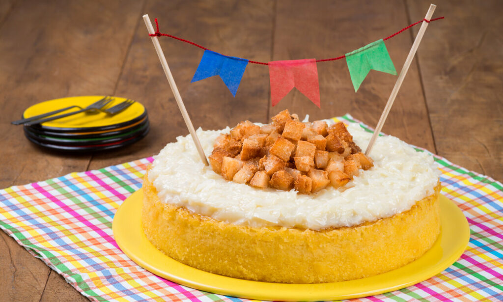 Bolo de milho redondo, coberto com calda de coco. Sobre a calda temos coquinhos doces. O bolo está em uma travessa redonda na cor amarela, disposta sobre uma toalha de mesa xadrex colorida. Ao lado pratos e garfos.
