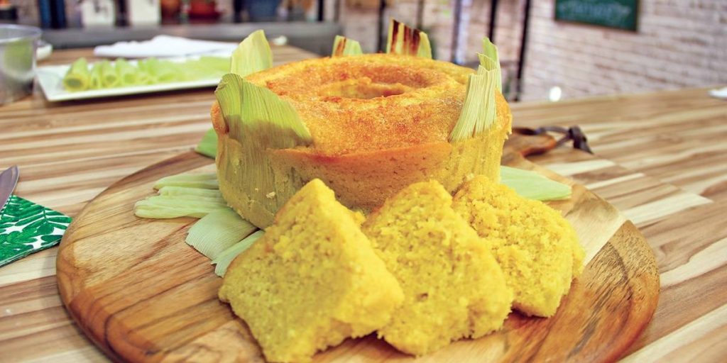 Bolo de milho inteiro já assado. Ele está decorado com algumas palhas e está sobre uma tábua de madeira redonda. Ao lado do bolo, algumas fatias.