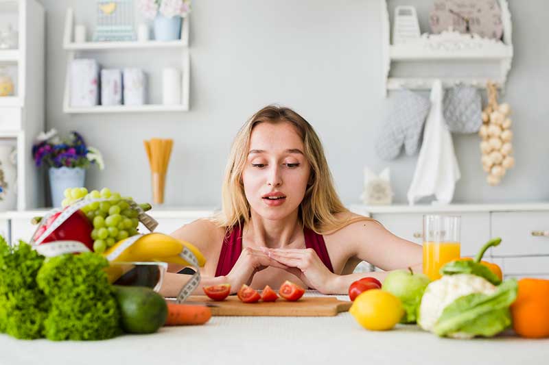 Mulher na cozinha. Ela está sentada de frente para um balcão e sobre ele temos vários tipos de frutas, tomate, alface, cenoura, couve flor, suco de laranja e uma fita métrica. A cozinha é clara e ao fundo uma parede com utensílios domésticos.