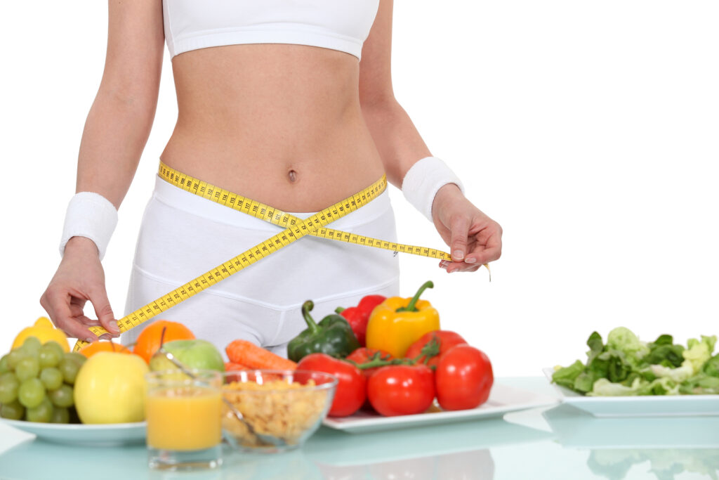Mulher com corpo escultural. Está de top, bermuda e munhequeira branca e usando uma fita métrica em torno de sua cintura. À sua frente, três pratos com frutas, legumes e verduras, um copo com suco de laranja.