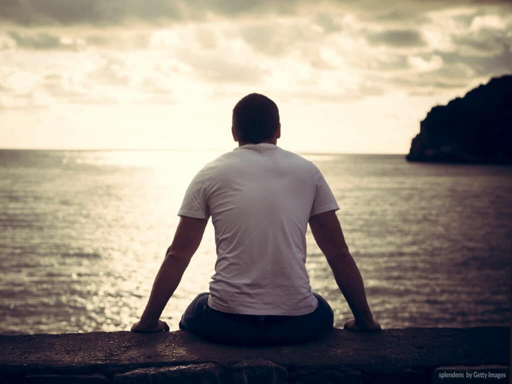 Homem sentado sobre uma muralha de pedra olhando para o infinito do mar, tendo ao fundo o por do sol. Ele está usando uma camiseta na cor branca e uma calça jeans.
