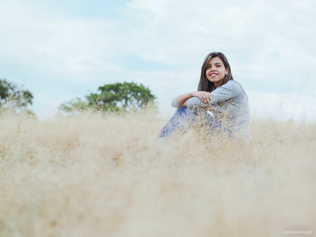 Mulher sorrindo em um campo de algodão. Ela está sentada e ao fundo da paisagem temos duas copas de árvores na cor verde.