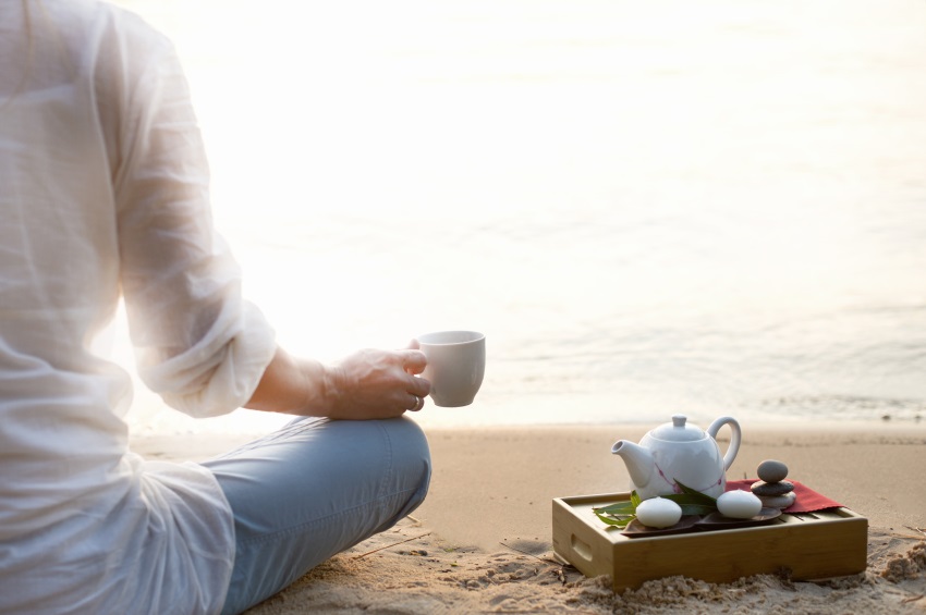 Imagem de metade do corpo de uma mulher. Ela veste uma camisa branca e calça azul clara. Está em posição de meditação, olhando para o mar. Em uma das mãos segura uma xícara de café. Ao seu lado, uma bandeja com um bule branco.