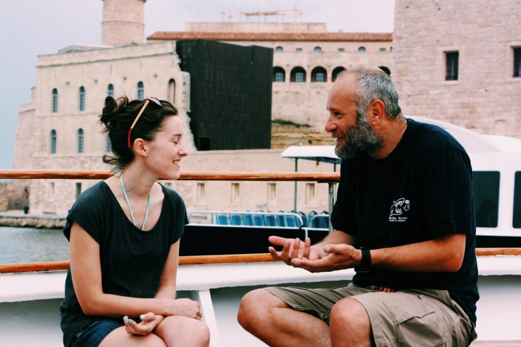 Um homem mais velho e uma mulher estão sentados em um barco, conversando em frente a um rio.