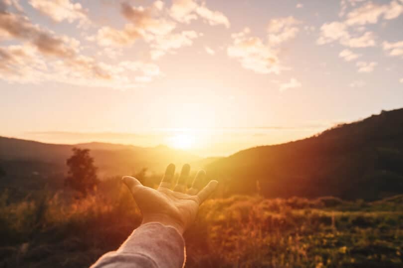 Mão de um homem espalmada para cima, em direção ao Sol que está se pondo.