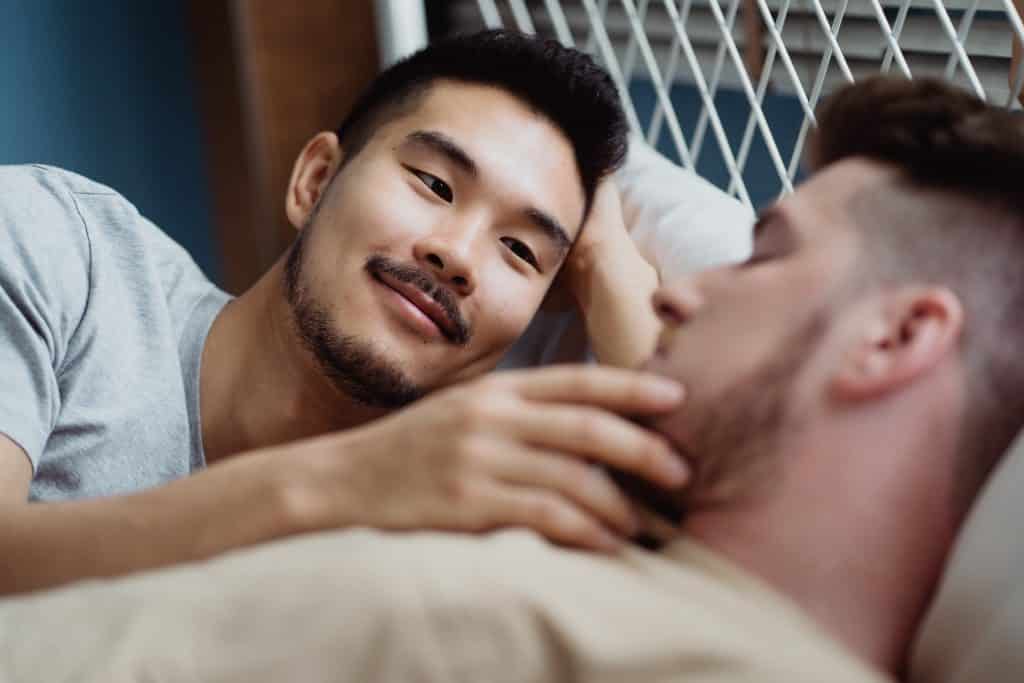 Dois homens deitados na cama, abraçados, enquanto um acaricia o rosto do outro.