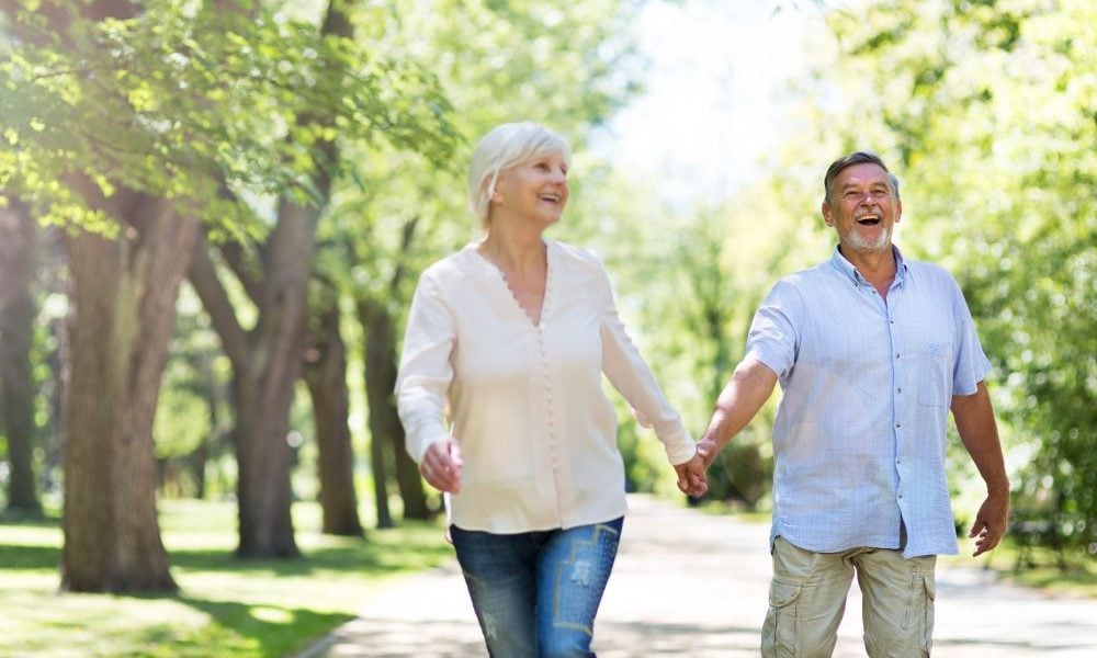 Casal de mãos dadas, caminhando em um parque cheio de árvores em um dia ensolarado. Ambos estão alegres e sorrindo.