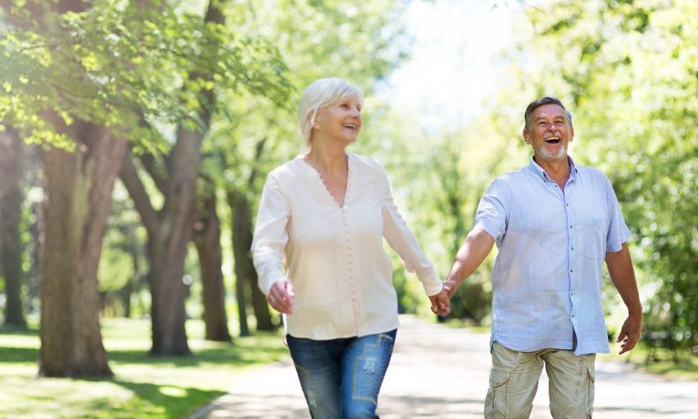 Casal de mãos dadas, caminhando em um parque cheio de árvores em um dia ensolarado. Ambos estão alegres e sorrindo.
