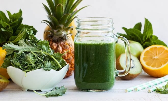 Copo de vidro com suco de couve, rodeado por outras frutas, como abacaxi e laranja.