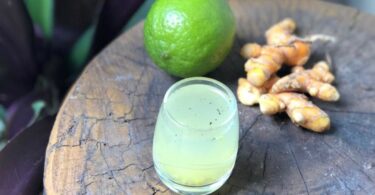 Copo com suco ao lado limão e gengibre sobre uma mesa de madeira.