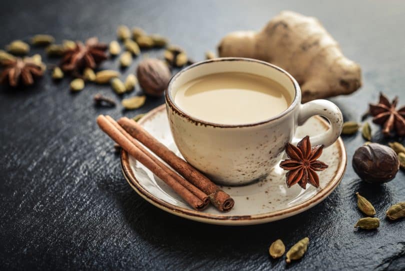 Caneca com chá de canela e gengibre. Na mesa há a caneca em um pires, uma lasca de canela e um pedaço de gengibre.