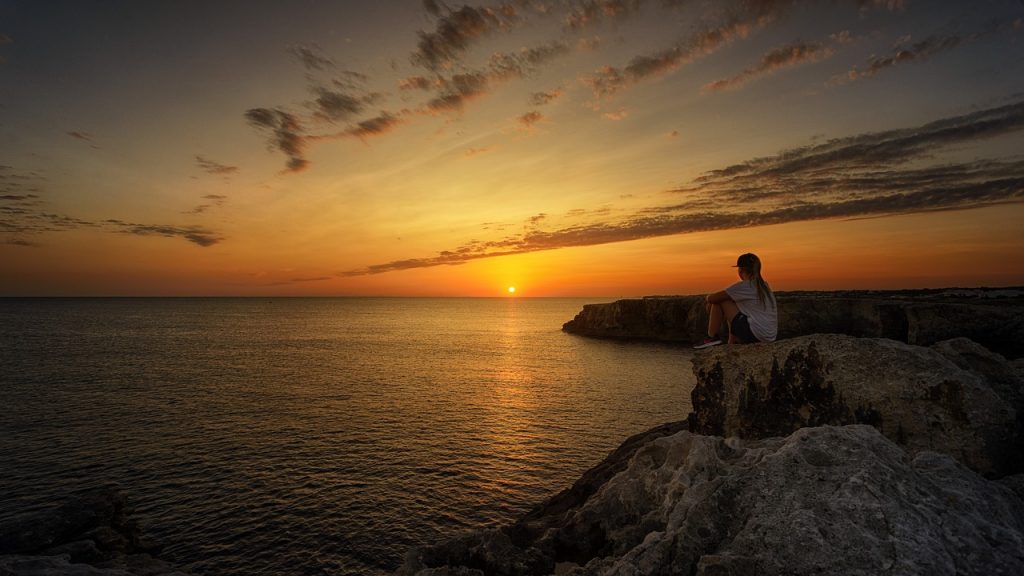 Imagem de um mar. Ao fundo o pôr do sol. Ao seu entorno várias pedras grandes. Em uma delas, encontra-se uma mulher sentada, olhando para o pôr do sol.