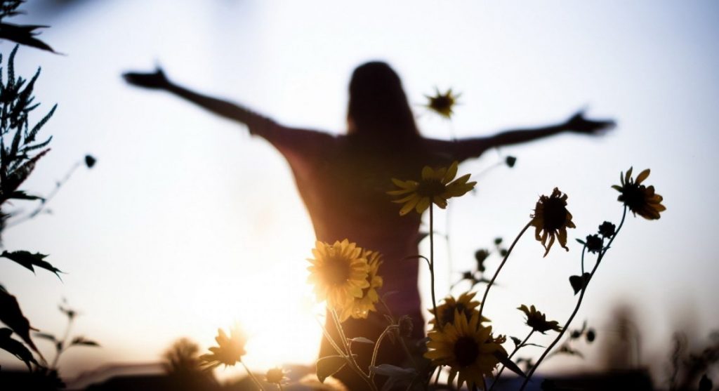 Mulher de braças abertos em um campo florido. Ela está de frente para o Sol. Em destaque na imagem temos margaridas na cor amarelo.