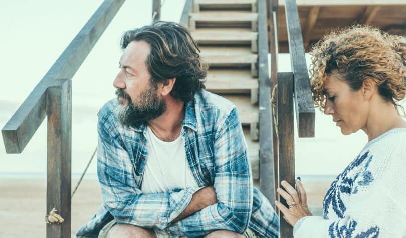 Casal em separação, com ar reflexivo. Ele está sentado em uma escada de madeira, na praia, olhando ao longe. Ela apoia a testa no corrimão da escada, ao lado dele, em desalento.