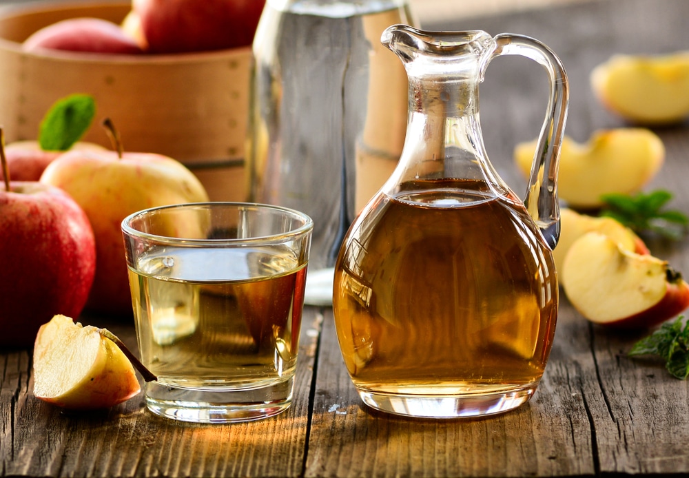 Uma jarra de vidro com vinagre de maçã. Ao lado dela um copo de vidro cheio de vinagre de maçã. Ambos estão dispostos sobre uma mesa de madeira com maças inteiras e cortadas ao meio.