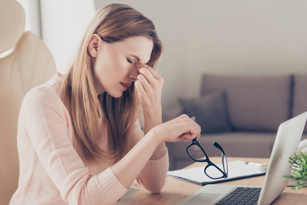 Mulher de cabelos longos, sentada em uma mesa no escritório de frente para o seu notebook. Ela está com dores de sinusite e segura em uma de suas mãos seus óculos.