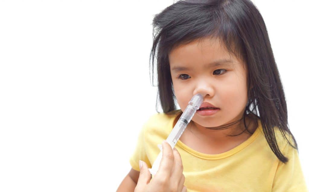Criança asiática vestindo camiseta amarela e sendo medicada com a aplicação de um remédio no seu nariz com o uso de uma seringa.
