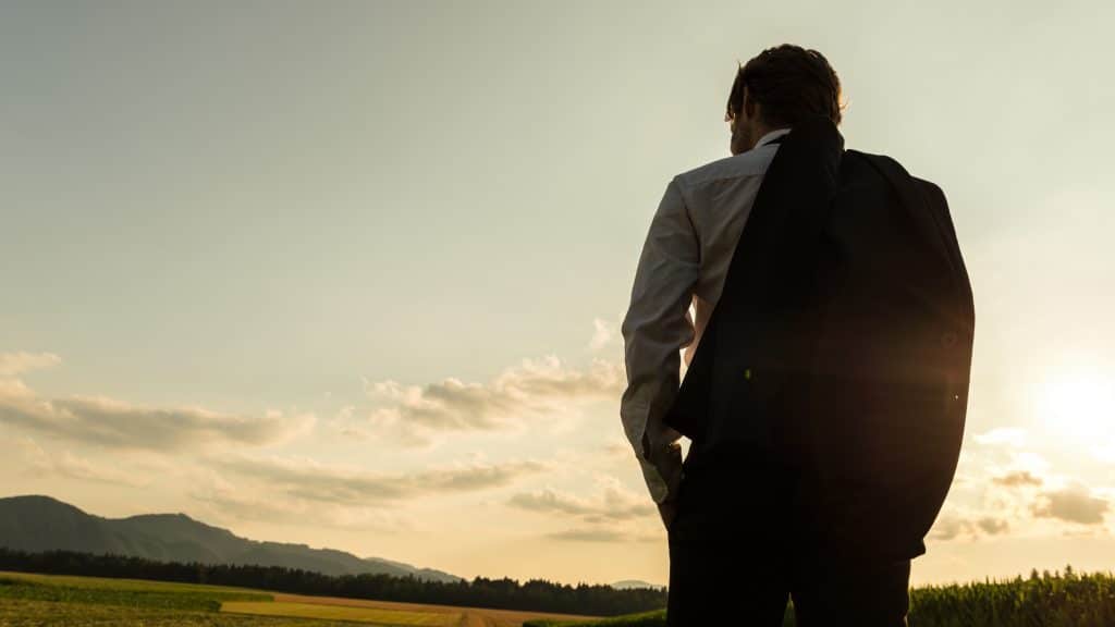 Homem de costas olhando para o horizonte com o sol se pondo. Ele está usando camisa branca e um terno pendurado no ombro.