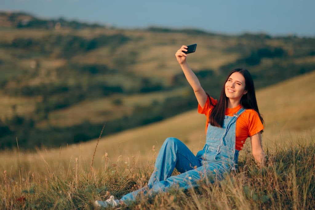 Uma mulher de cabelos longos, vestida de macacão jeans claro e camiseta laranja. Ela está tirando uma selfie em meio a natureza.