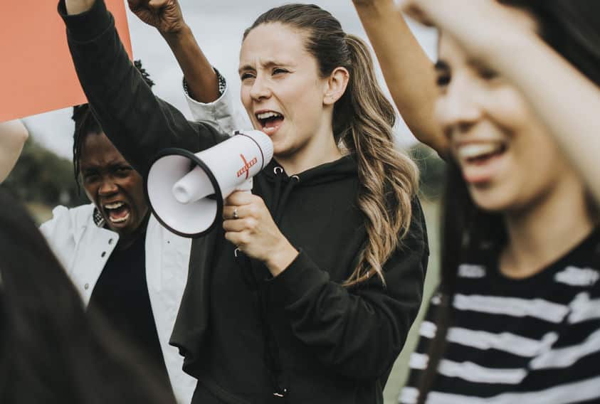 Grupo de mulheres protestando. Uma delas segura um cartaz, e outra fala em um megafone.