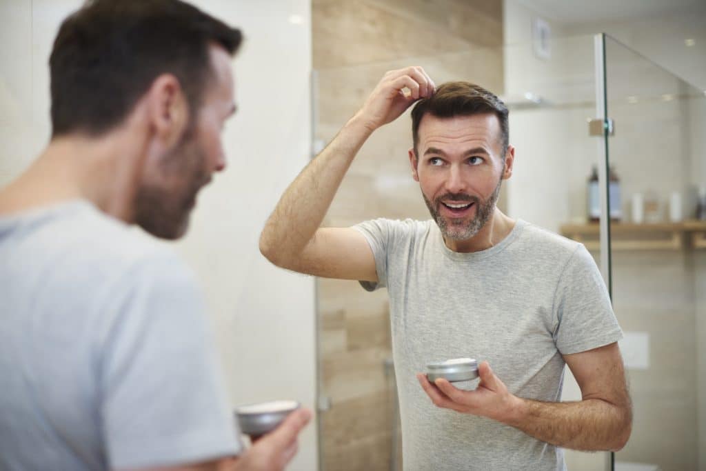 Imagem de um homem refletida no espelho de um banheiro. Ele está cuidado de sua pele e de seu cabelo.
