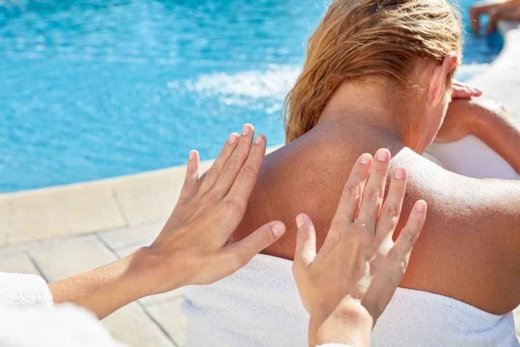 Mulher próxima a uma piscina. Ela está sentada e enrolada em uma toalha branca recebendo massagem na região das costas e nuca.