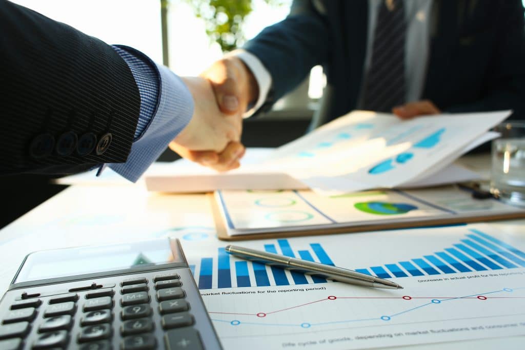 Dois homens de negócios dando as mãos em forma de agradecimento pelos resultados financeiros obtidos. Eles estão sentados frente a uma mesa com vários relatórios financeiros, calculadora e caneta espalhados.
