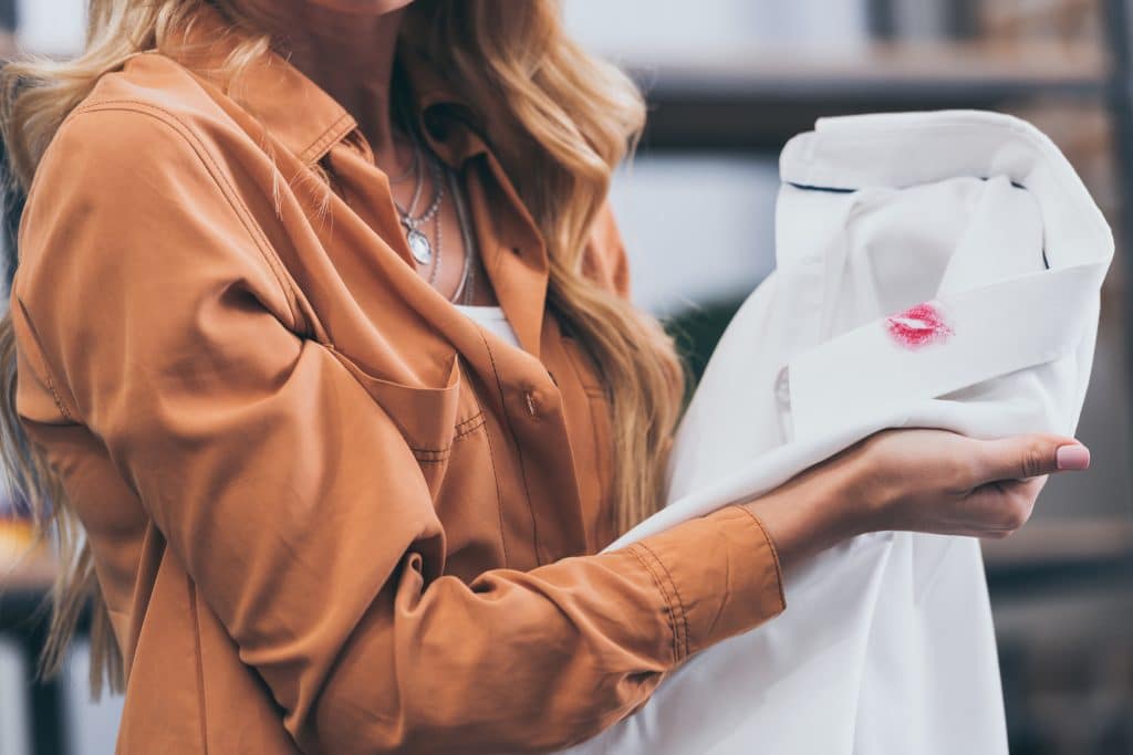 Mulher vestida com uma camisa marrom. Ela está segurando a camisa branca do esposo. No colarinho da camisa tem uma mancha de batom vermelho.