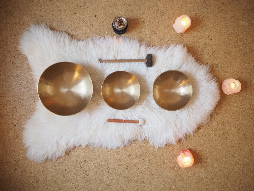Vários símbolos que representam o budismo tibetano. Na imagem temos: taças tibetanas, castiçais de velas acesas, tapete branco de pelo. Tudo para promover o som de cura, meditação e relaxamento.