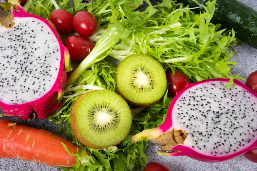 Pitaya cortada ao meio. Ao lado dela temos uma cenoura, tomates cereja, um kiwi também cortado ao meio e algumas folhas de alface.
