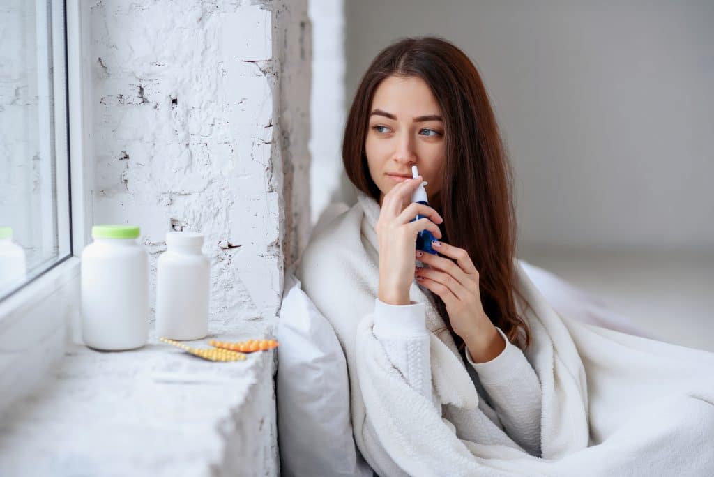 Mulher em seu quarto. Ela está sentada em sua cama. Veste um pijama branca e usa uma coberta branca. Ela está usando um spray nasal.