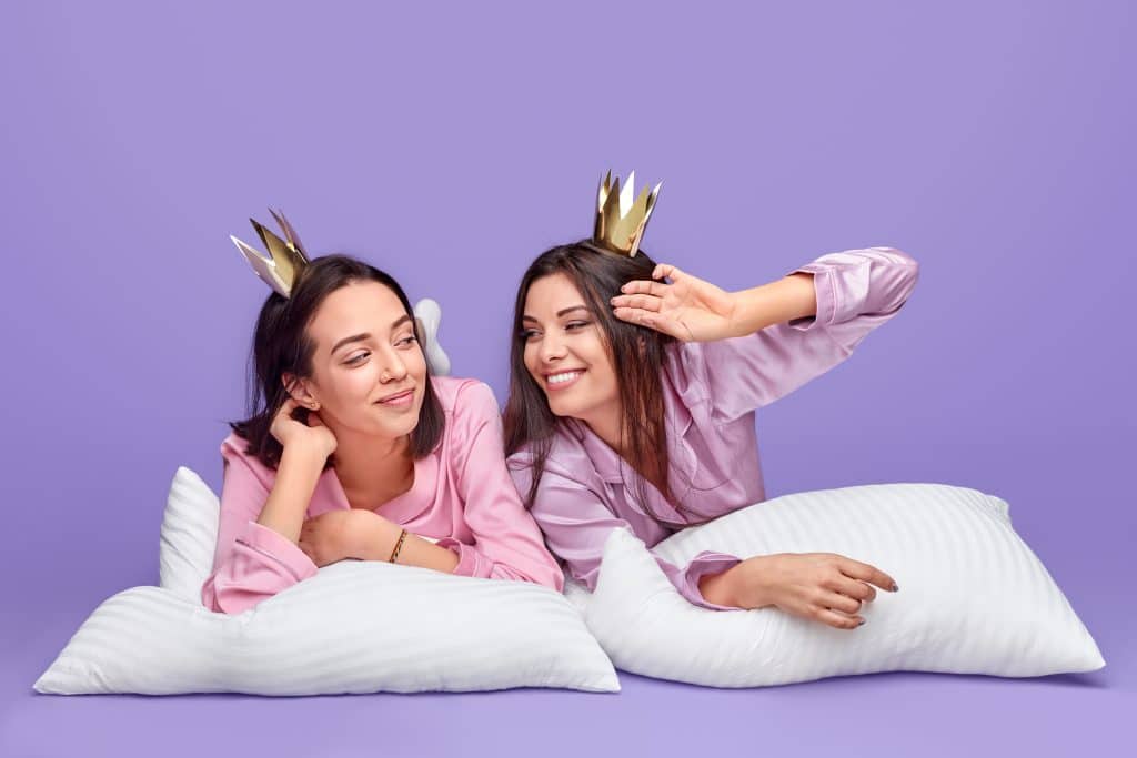 Duas mulheres de bruço em dois travesseiros brancos, sendo uma mulher em cada um. Elas vestem um pijama rosa e usam coroas douradas.