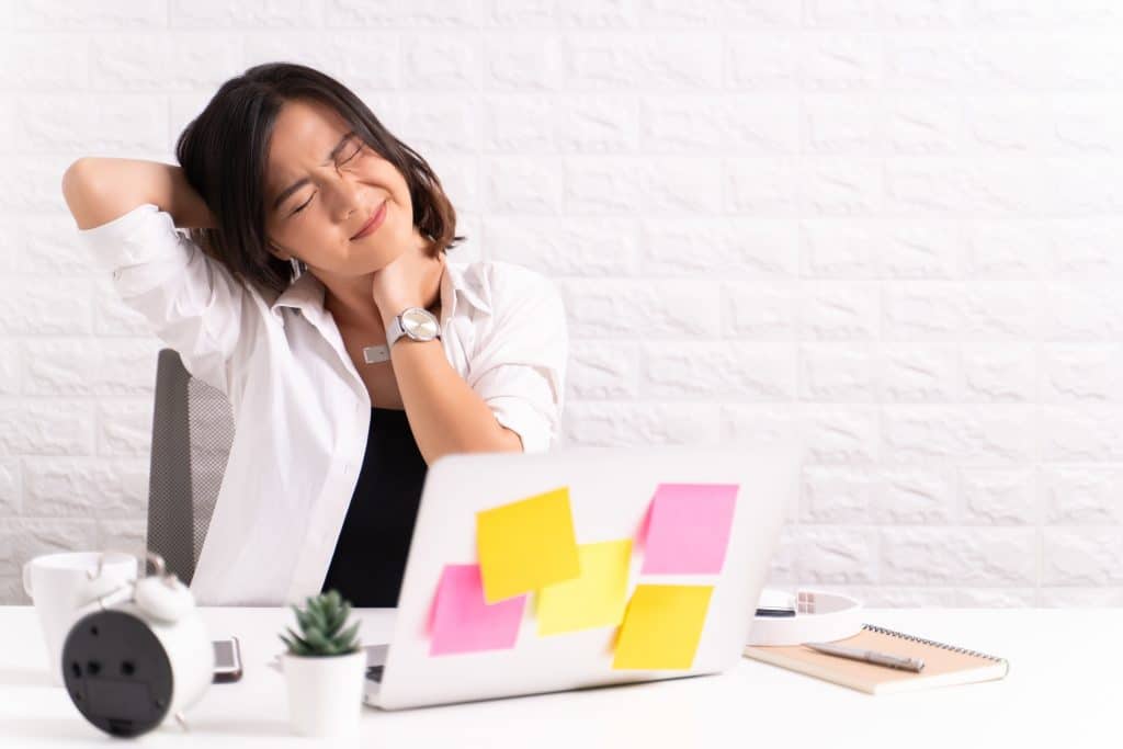 Mulher em sua sala de escritório com paredes e decoração branca. Ela está de frente para o notebook cheio de post-it colorido. Ela está com dores na nuca, pois suas mãos estão sobre ela.