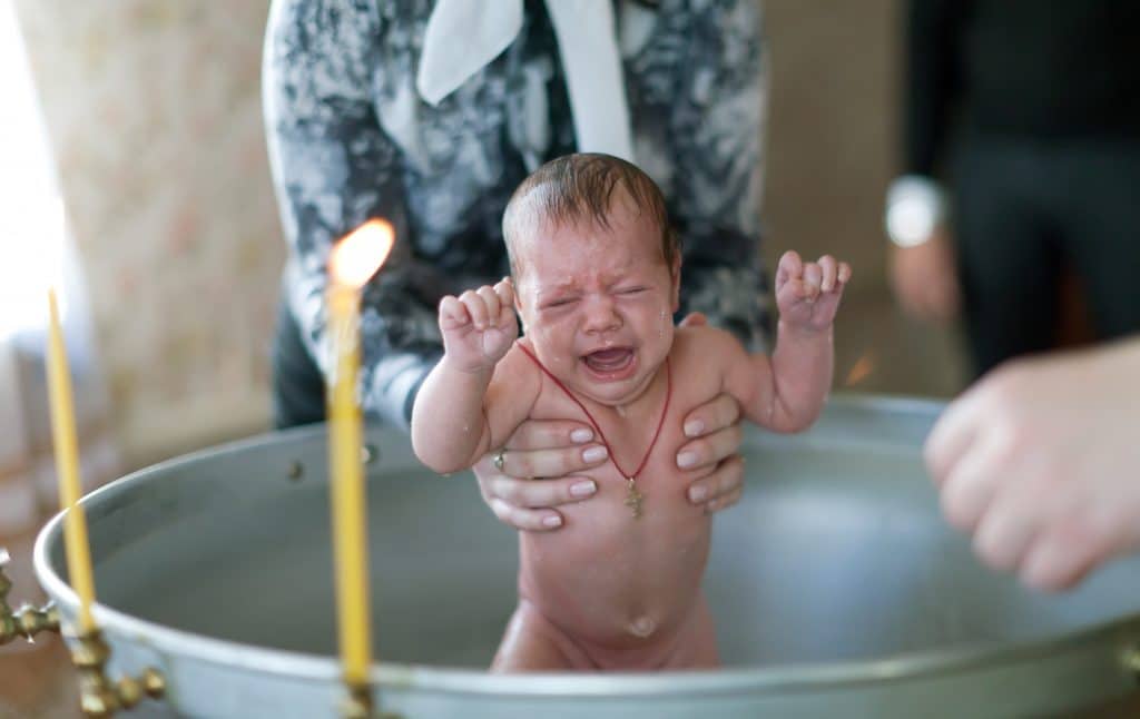 Criança chorando dentro de um tanque de batismo. Ao redor do tanque velas acesas. Uma mulher segura a criança pelos braços.
