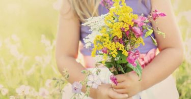 Menina usando vestido segurando um buque de flores em uma campina. A foto corta sua cabeça, vemos apenas seu corpo.