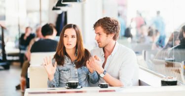 Casal sentado lado a lado numa lanchonete. O homem olha para a mulher e segura sua mão esquerda. A mulher olha para o outro lado, e a mão direita levantada.