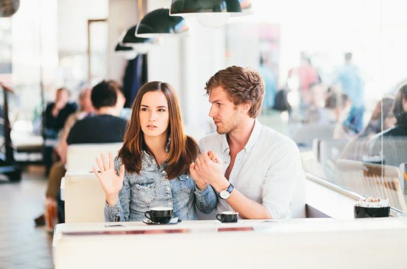 Casal sentado lado a lado numa lanchonete. O homem olha para a mulher e segura sua mão esquerda. A mulher olha para o outro lado, e a mão direita levantada.