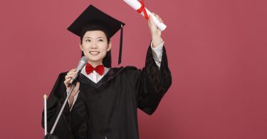 Mulher vestindo uma beca de formatura segura um diploma enquanto fala ao microfone.