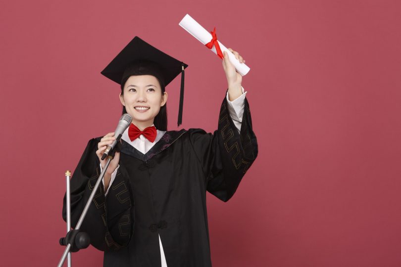 Mulher vestindo uma beca de formatura segura um diploma enquanto fala ao microfone.