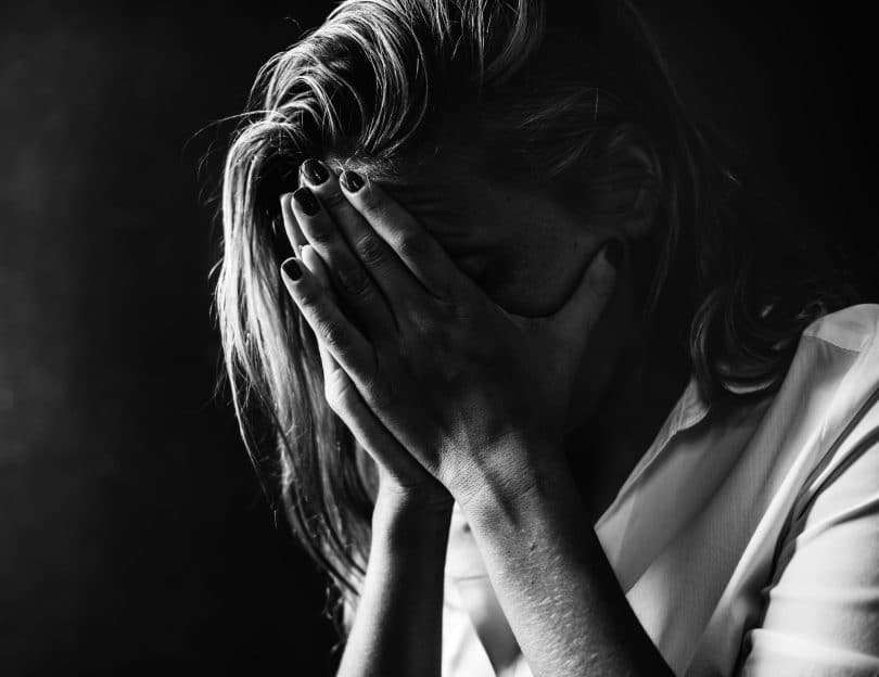 Foto em preto e branco de uma mulher triste com as mãos escondendo o rosto.