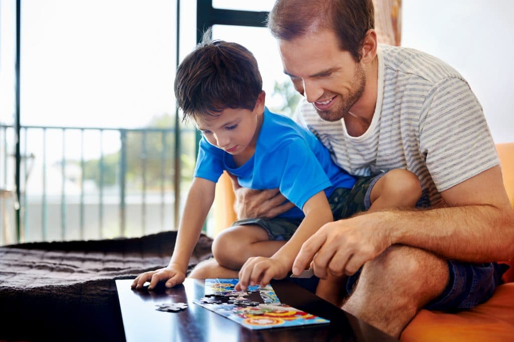 Pai e filho montando um quebra-cabeça. Eles estão na sala de estar, sentados em um sofá laranja.