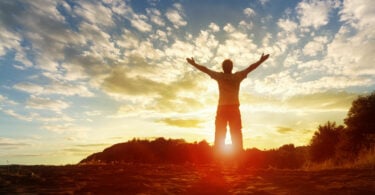 Silhueta de um homem com as mãos levantadas com por do sol e céu com nuvens ao fundo