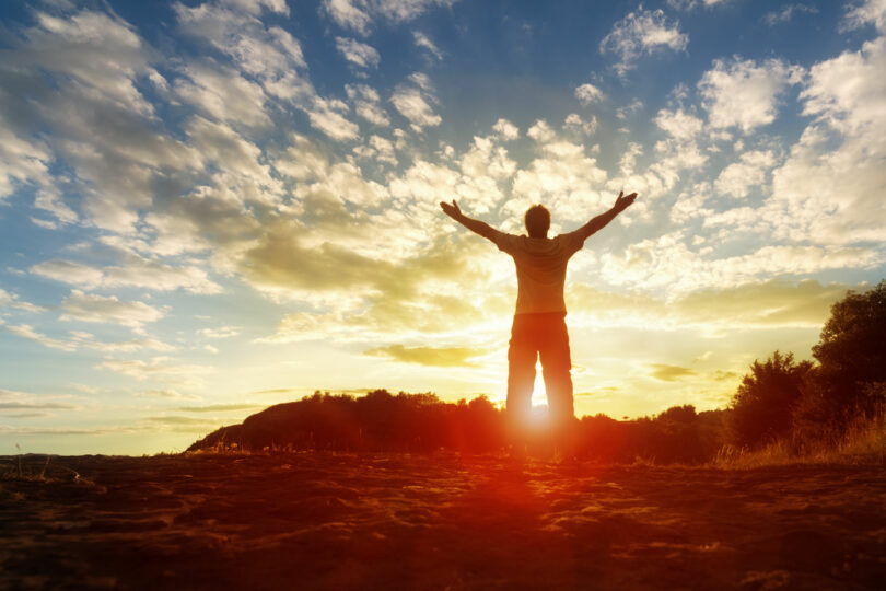 Silhueta de um homem com as mãos levantadas com por do sol e céu com nuvens ao fundo