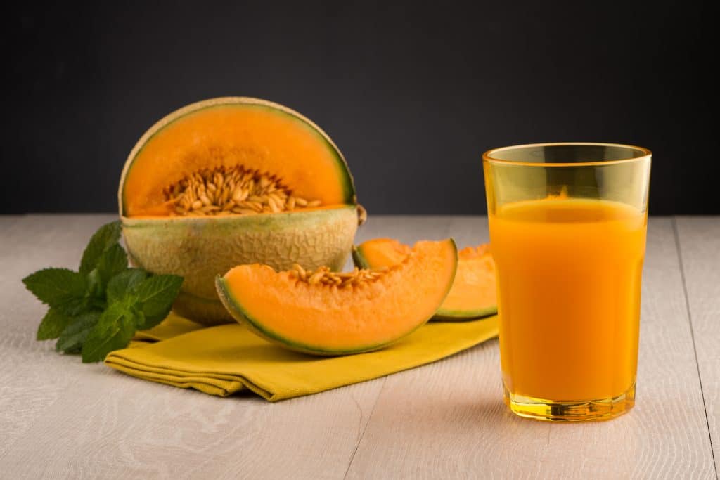 Suco de melão e hortelã para a gordura no fígado. Ele está sendo servido em um copo de vidro sobre a mesa de madeira. Ao lado um melão cortado e duas fatias da fruta com algumas folhas de hortelã.